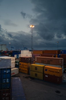 Cargo containers stacked in a port setting under a cloudy sky. Industrial cranes are visible in the background, and a tall light post illuminates the area.