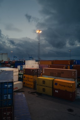 Cargo containers stacked in a port setting under a cloudy sky. Industrial cranes are visible in the background, and a tall light post illuminates the area.