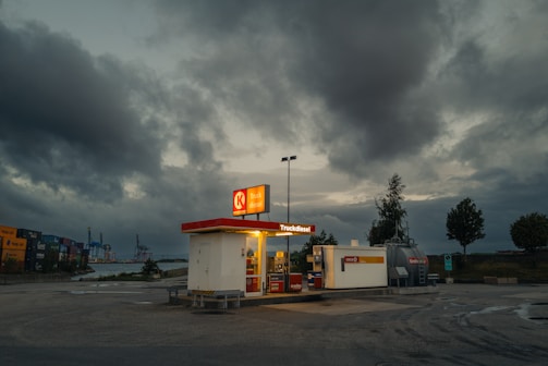A solitary diesel gas station stands under a dramatic, overcast sky, suggesting an industrial setting. The lighting from the station contrasts with the dark clouds above, and a few trees are visible in the background. Cargo containers and industrial cranes can be seen in the distance, highlighting a dockyard ambiance.