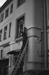 A person is standing on a ladder leaning against a building facade, appearing to perform maintenance or repairs on a window or the wall. The building has several windows and an entrance canopy. The image is in black and white, giving it a classic, timeless feel.