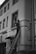 A person is standing on a ladder leaning against a building facade, appearing to perform maintenance or repairs on a window or the wall. The building has several windows and an entrance canopy. The image is in black and white, giving it a classic, timeless feel.