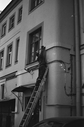 A person is standing on a ladder leaning against a building facade, appearing to perform maintenance or repairs on a window or the wall. The building has several windows and an entrance canopy. The image is in black and white, giving it a classic, timeless feel.