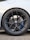 A close-up view of a car wheel featuring a detailed design with a black alloy rim. The tire is a Michelin Pilot Sport with visible branding. The wheel includes a ventilated disc brake with blue calipers and the brand logo at the center of the hub. The car body is white and the ground is paved with small stones.