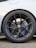 A close-up view of a car wheel featuring a detailed design with a black alloy rim. The tire is a Michelin Pilot Sport with visible branding. The wheel includes a ventilated disc brake with blue calipers and the brand logo at the center of the hub. The car body is white and the ground is paved with small stones.