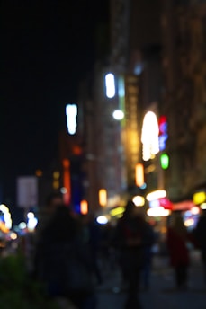 A blurry image of a city street at night, featuring bright lights and indistinct silhouettes of people walking. Neon signs and illuminated structures create a vibrant but unfocused scene, emphasizing the bustling atmosphere of an urban nightlife.