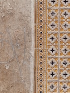 A close-up view of a textured wall with a combination of plain, rough plaster and decorative ceramic tiles. The left side features a faded beige surface with visible cracks and weathering. The right side showcases ornate tiles in a repeating pattern with yellow and black floral motifs, bordered by a column of smaller, circular designs.