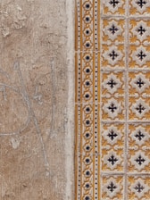 A close-up view of a textured wall with a combination of plain, rough plaster and decorative ceramic tiles. The left side features a faded beige surface with visible cracks and weathering. The right side showcases ornate tiles in a repeating pattern with yellow and black floral motifs, bordered by a column of smaller, circular designs.