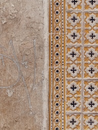 A close-up view of a textured wall with a combination of plain, rough plaster and decorative ceramic tiles. The left side features a faded beige surface with visible cracks and weathering. The right side showcases ornate tiles in a repeating pattern with yellow and black floral motifs, bordered by a column of smaller, circular designs.