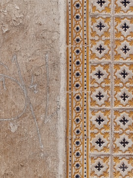 A close-up view of a textured wall with a combination of plain, rough plaster and decorative ceramic tiles. The left side features a faded beige surface with visible cracks and weathering. The right side showcases ornate tiles in a repeating pattern with yellow and black floral motifs, bordered by a column of smaller, circular designs.