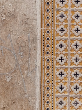 A close-up view of a textured wall with a combination of plain, rough plaster and decorative ceramic tiles. The left side features a faded beige surface with visible cracks and weathering. The right side showcases ornate tiles in a repeating pattern with yellow and black floral motifs, bordered by a column of smaller, circular designs.