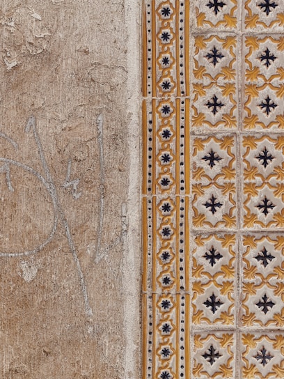 A close-up view of a textured wall with a combination of plain, rough plaster and decorative ceramic tiles. The left side features a faded beige surface with visible cracks and weathering. The right side showcases ornate tiles in a repeating pattern with yellow and black floral motifs, bordered by a column of smaller, circular designs.
