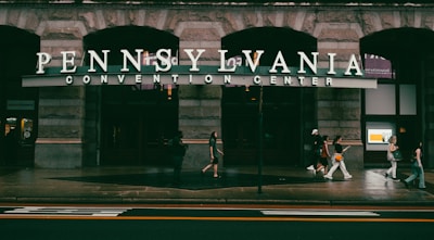 People walk past the entrance of the Pennsylvania Convention Center. The building features large stone arches and the entrance is marked by a prominent sign. The scene appears to be set in an urban environment with a sidewalk and a street in the foreground.