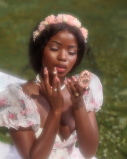A woman is adorned with a floral crown and is wearing a dress with floral patterns. She is gazing at a compact mirror while touching her face, surrounded by soft, natural lighting.