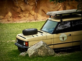 A partially buried safari vehicle is positioned in a grassy area near large rocks. The vehicle has a brownish-yellow color, with a spare tire attached to the front hood and a roof rack on top. It displays a logo for a park service on its door. In the background, a textured, rocky wall with patches of grass beneath can be seen.