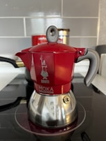 A red and silver stovetop coffee maker, known as a moka pot, is placed on an induction cooktop. The pot features a shiny bottom with a distinct handle and a spout, along with a visible brand logo and text on its body. The background includes a tiled wall and a partially visible container.