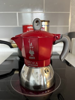 A red and silver stovetop coffee maker, known as a moka pot, is placed on an induction cooktop. The pot features a shiny bottom with a distinct handle and a spout, along with a visible brand logo and text on its body. The background includes a tiled wall and a partially visible container.