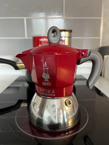 A red and silver stovetop coffee maker, known as a moka pot, is placed on an induction cooktop. The pot features a shiny bottom with a distinct handle and a spout, along with a visible brand logo and text on its body. The background includes a tiled wall and a partially visible container.