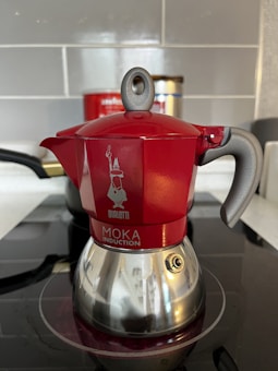 A red and silver stovetop coffee maker, known as a moka pot, is placed on an induction cooktop. The pot features a shiny bottom with a distinct handle and a spout, along with a visible brand logo and text on its body. The background includes a tiled wall and a partially visible container.