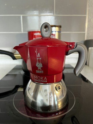A red and silver stovetop coffee maker, known as a moka pot, is placed on an induction cooktop. The pot features a shiny bottom with a distinct handle and a spout, along with a visible brand logo and text on its body. The background includes a tiled wall and a partially visible container.