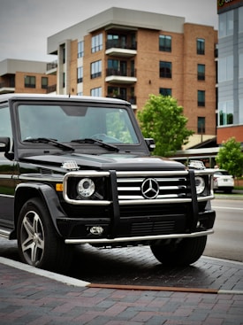 A black luxury SUV, specifically a Mercedes-Benz G-Class, is parked on a brick pavement in an urban setting. The background features modern apartment buildings with red brick and beige facades, as well as some green trees. The scene appears to be a calm neighborhood area.