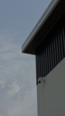 A security camera is mounted on the corner of an industrial-style building with corrugated metal siding. The view focuses on the building's geometric lines, contrasting with a cloudy sky in the background.