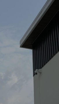 A security camera is mounted on the corner of an industrial-style building with corrugated metal siding. The view focuses on the building's geometric lines, contrasting with a cloudy sky in the background.