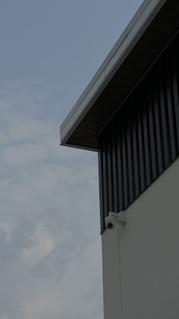 A security camera is mounted on the corner of an industrial-style building with corrugated metal siding. The view focuses on the building's geometric lines, contrasting with a cloudy sky in the background.
