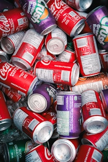 A collection of various soda cans with vibrant colors, including brands like Coca-Cola and Crush, mingled together with visible condensation and scattered ice cubes.