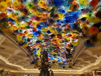A stunning ceiling installation featuring a multitude of colorful glass flowers in various shapes and sizes. The floral elements create a vibrant and dynamic display that resembles a blooming garden. The setting is a grand indoor space with elegant architectural details and soft lighting.