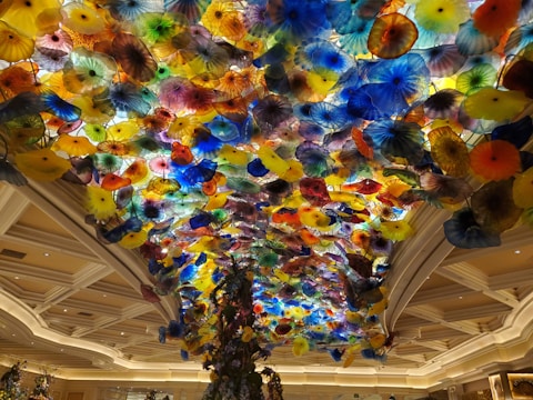 A stunning ceiling installation featuring a multitude of colorful glass flowers in various shapes and sizes. The floral elements create a vibrant and dynamic display that resembles a blooming garden. The setting is a grand indoor space with elegant architectural details and soft lighting.