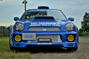 A Subaru WRX car is parked on a gravel path. The car features a vibrant blue exterior with yellow fog lights and a prominent hood scoop. It has decals for the Subaru World Rally Team and an Ontario license plate.