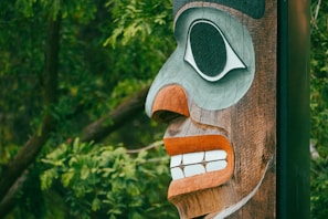 A detailed, carved wooden totem pole face set against a backdrop of vibrant green foliage. The carving features prominent eye and mouth designs, with contrasting colors of black, white, and orange.