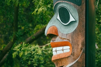 A detailed, carved wooden totem pole face set against a backdrop of vibrant green foliage. The carving features prominent eye and mouth designs, with contrasting colors of black, white, and orange.