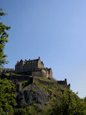 An ancient stone château perched atop a lush green hill under a clear blue sky.