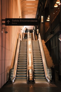 A modern indoor setting featuring two parallel escalators leading upwards, illuminated by soft lights. The walls have a sleek, tiled design and there is a sign indicating 'Salida Encomenderos' with 'Exit to'. The overall atmosphere is calm and structured.