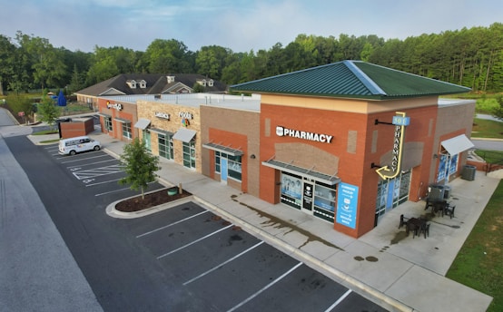 A small commercial plaza featuring a pharmacy and other shops. The brick buildings have modern signage and awnings. A few parked cars are visible, and there are outdoor seating areas. The surrounding area is landscaped with trees and grass.