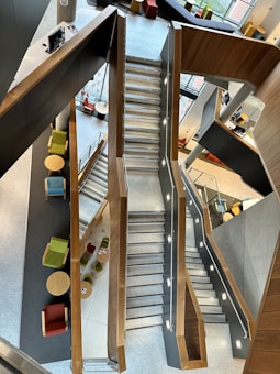 A modern, multi-level staircase with wooden railings and grey steps is depicted. Comfortable seating areas with colorful chairs and round tables surround the staircase, suggesting a communal space. The floor and walls have a sleek, contemporary design, with large windows providing natural light.