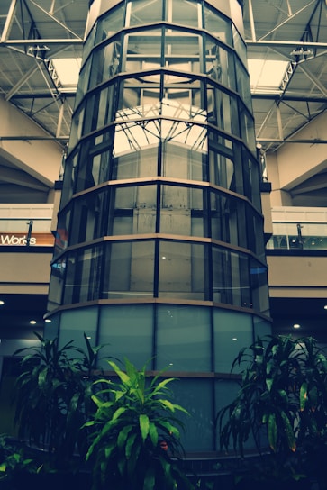 A tall, cylindrical glass structure surrounded by several floors in what appears to be an indoor commercial space. The bottom level is decorated with green plants, while metal beams and glass railings are visible on the upper floors. Light is filtering through the large ceiling windows.