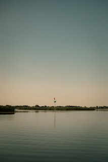 A calm body of water with a lone navigational marker standing in the middle. The sky has a muted gradient from blue to light yellow, and the horizon features distant, low-lying trees and vegetation.
