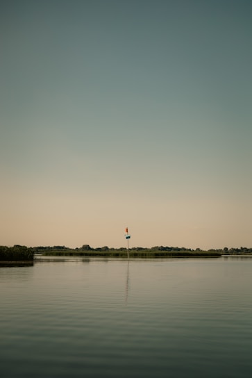 A calm body of water with a lone navigational marker standing in the middle. The sky has a muted gradient from blue to light yellow, and the horizon features distant, low-lying trees and vegetation.