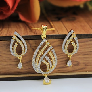 A gold pendant with matching earrings, intricately designed with teardrop shapes adorned with small, sparkling stones. The jewelry set rests against a wooden background, with delicate chains and clasps visible. The shine and elegance of the pieces suggest they are crafted for special occasions.