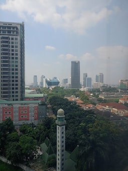 A cityscape featuring a tall skyscraper with numerous windows on the left and a modern high-rise building in the background, surrounded by smaller structures and lush greenery. A prominent mosque with a green roof and a dome-topped minaret stands out among the buildings, adding cultural significance to the scene. The sky is mostly clear with a few scattered clouds.