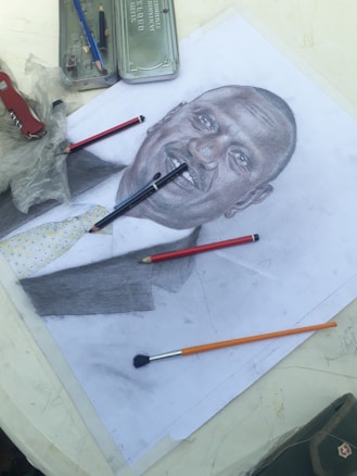 A detailed pencil sketch of a person is placed on a table, surrounded by art supplies including colored pencils, a paintbrush, and a metal pencil case. The sketch depicts the person in a suit and tie, with a focus on facial features and shading. The table has a light-colored surface.