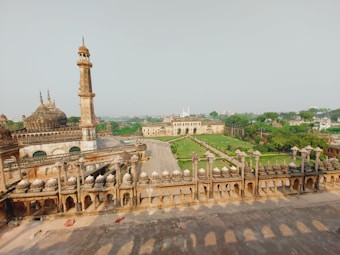 An expansive view of a historical architectural site featuring intricate designs and domes. A tall minaret stands prominently, surrounded by lush green gardens and pathways. There are structures with ornate railings and arches, indicating the cultural and historical significance of the site.