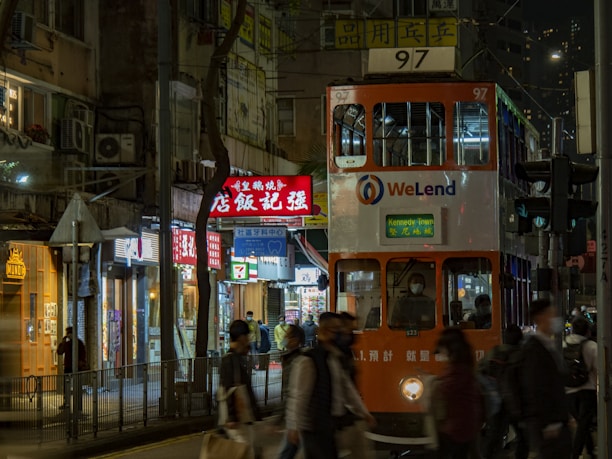 A bustling urban street scene featuring a double-decker tram with the number 97 heading towards Kennedy Town. The tram, adorned with the WeLend logo, moves through a street lined with brightly lit shop signs in various languages. Several pedestrians, some wearing masks, walk past the tram and shops.