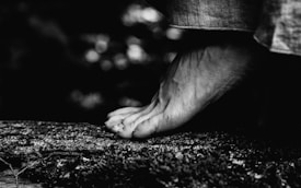 A monochrome image captures the delicate interplay of form and texture, featuring a close-up of a foot poised gently on a rough, textured surface, possibly a log. The details of the skin and the denim fabric are accentuated against a softly blurred background.