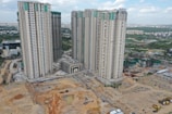 Multiple high-rise residential buildings under construction are surrounded by a mix of cleared land and other construction activities. The area is partly developed with a combination of dirt and concrete surfaces. Nearby, several smaller buildings and construction equipment can be seen, with green netting on some structures indicating an ongoing project.