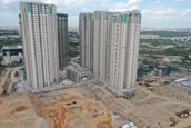 Multiple high-rise residential buildings under construction are surrounded by a mix of cleared land and other construction activities. The area is partly developed with a combination of dirt and concrete surfaces. Nearby, several smaller buildings and construction equipment can be seen, with green netting on some structures indicating an ongoing project.