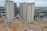 Multiple high-rise residential buildings under construction are surrounded by a mix of cleared land and other construction activities. The area is partly developed with a combination of dirt and concrete surfaces. Nearby, several smaller buildings and construction equipment can be seen, with green netting on some structures indicating an ongoing project.