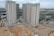 Multiple high-rise residential buildings under construction are surrounded by a mix of cleared land and other construction activities. The area is partly developed with a combination of dirt and concrete surfaces. Nearby, several smaller buildings and construction equipment can be seen, with green netting on some structures indicating an ongoing project.